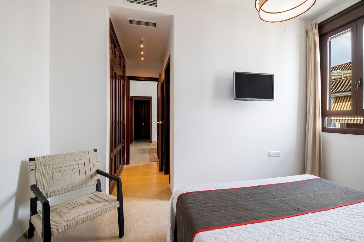A hotel room with a bed, a television mounted on the wall, a chair, and a window with curtains.