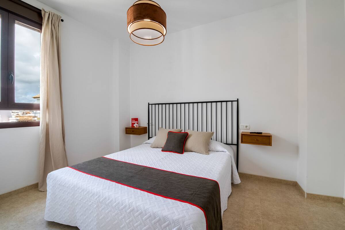 A neatly made bed with a white and gray bedspread, a red-lined headboard, and a bedside table with a lamp.