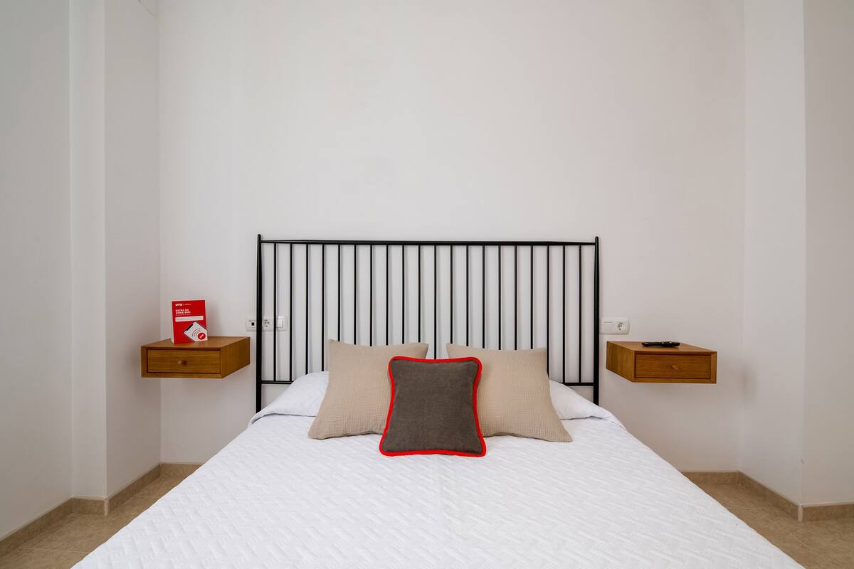A neatly made bed with a white comforter, two beige pillows, and a gray pillow with a red trim. There are two wooden nightstands, each with a red box and a remote control.
