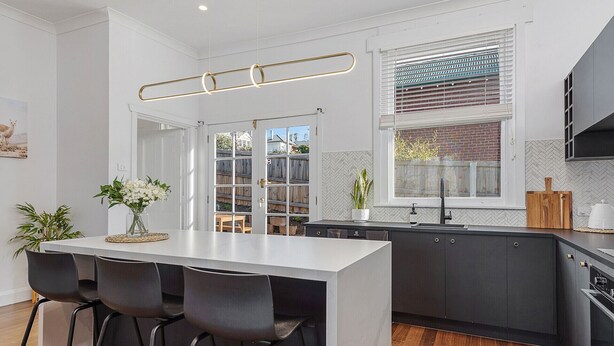 A modern kitchen with a central island, black bar stools, a large window with blinds, and a herringbone-patterned backsplash.