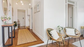 A hallway with a wooden floor, a rug, wicker chairs with white cushions, a mirror, and a hanging light fixture.