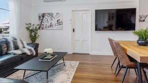 A modern living room with a black leather sofa, a wooden dining table with chairs, a flat-screen TV, and artwork on the walls.