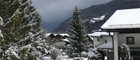 A snow-covered landscape with a mountain in the background, a pathway leading to a building with a snow-covered roof, and snow-laden trees.