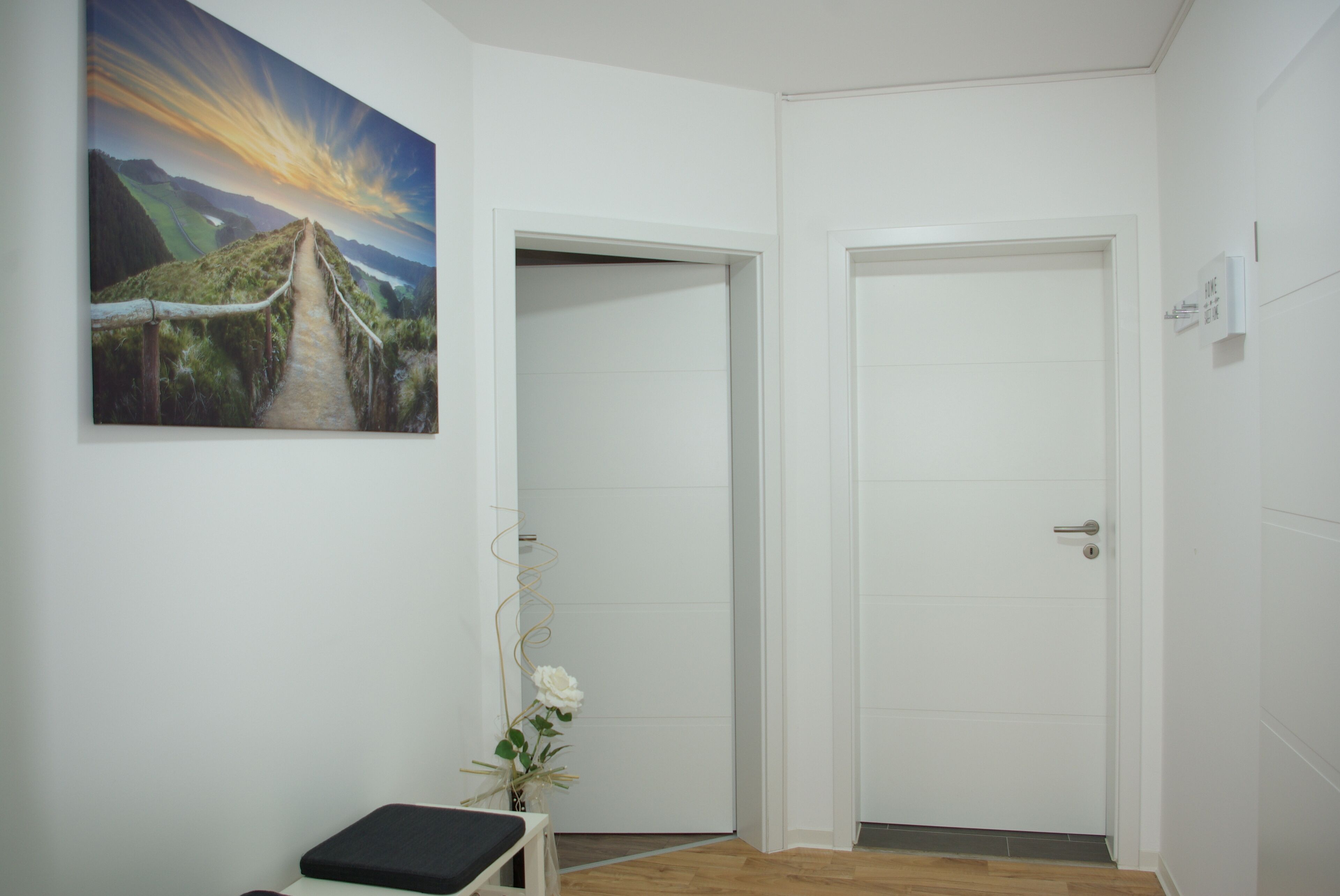 Een kamer met een houten vloer, witte muren, een landschapsschilderij, een witte deur en een kleine tafel met een bloevas.