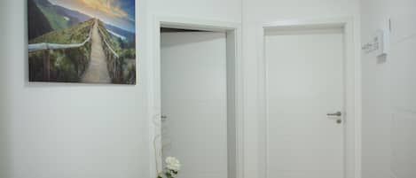 Een kamer met een houten vloer, witte muren, een landschapsschilderij, een witte deur en een kleine tafel met een bloevas.