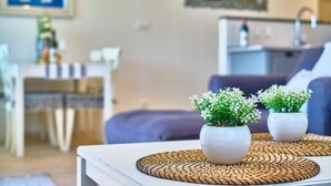 Un salón moderno con un sofá blanco, dos plantas en macetas sobre un tapete tejido, una cocina al fondo y un cuadro en la pared.