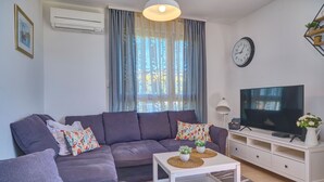 Una sala de estar moderna con un sofá gris, una mesa de centro blanca con dos plantas en macetas, un televisor de pantalla plana sobre un mueble blanco y un ventanal con cortinas translúcidas.