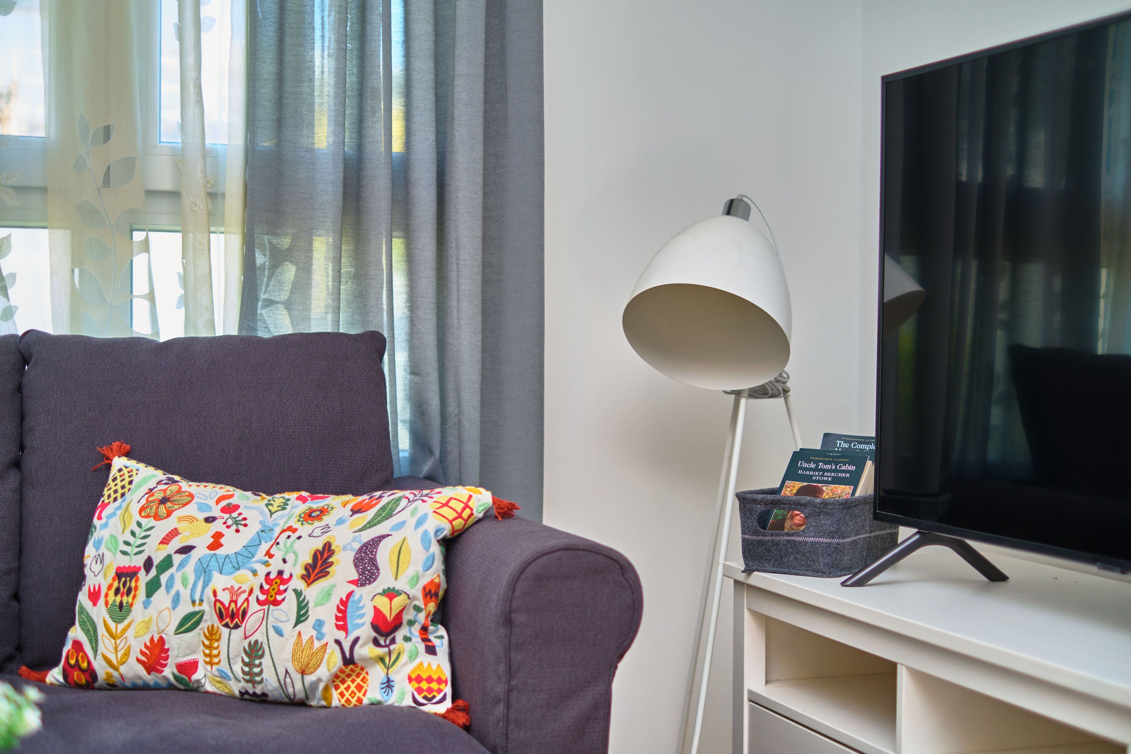 Un saló amb un sofà gris, coixins florals de colors, una làmpada de peu blanca i un televisor de pantalla plana sobre un suport blanc.