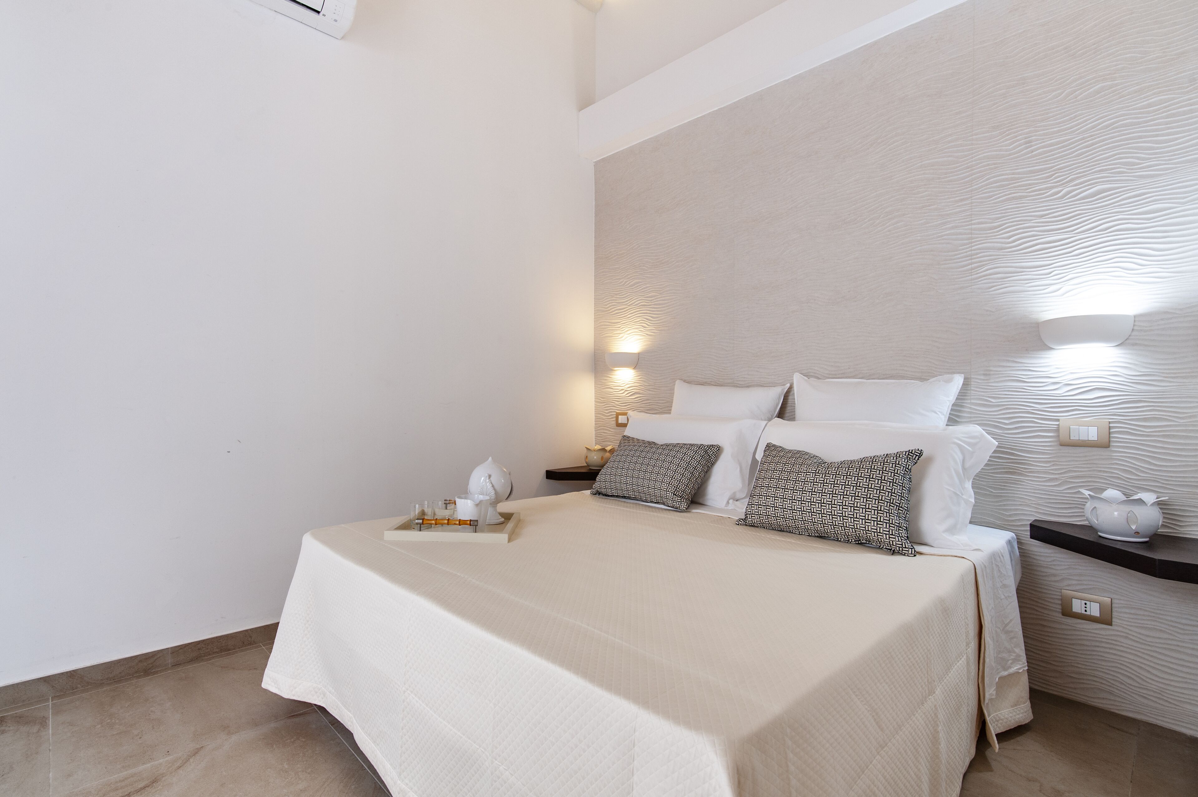 Una cama bien hecha con una colcha blanca, dos almohadas y una mesita de noche con una lámpara y un pequeño florero.