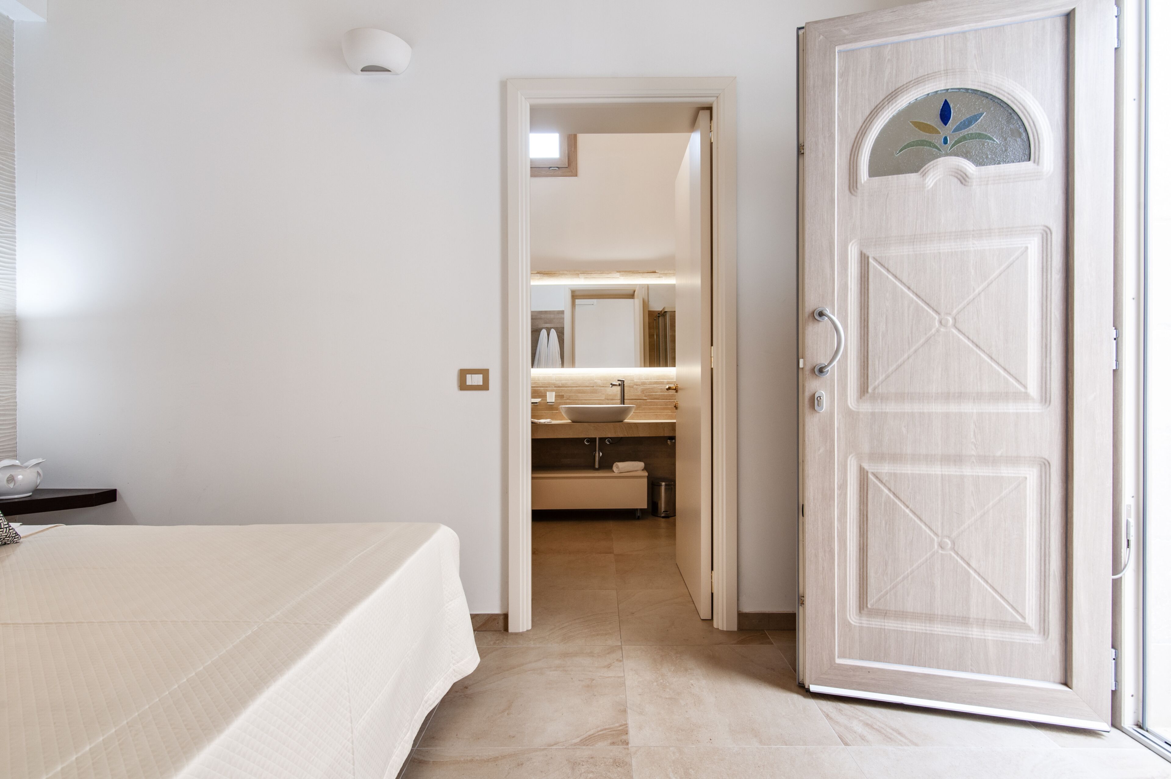 A hotel room with a bed, a bedside table, a bathroom with a sink and mirror, and a door with a decorative glass panel.