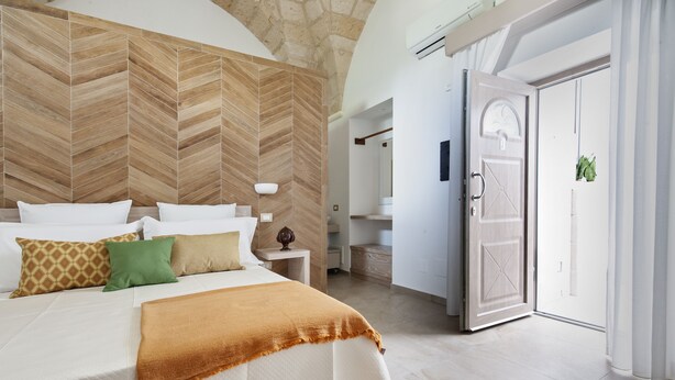 Un dormitorio con una cama grande, un revestimiento de madera en espiga, una mesita de noche con lámpara y una puerta que da a otra habitación.
