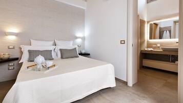 Una habitación de hotel moderna con una cama grande, lámparas de noche, una mesita con un vaso y una taza, y un baño con lavabo y espejo.