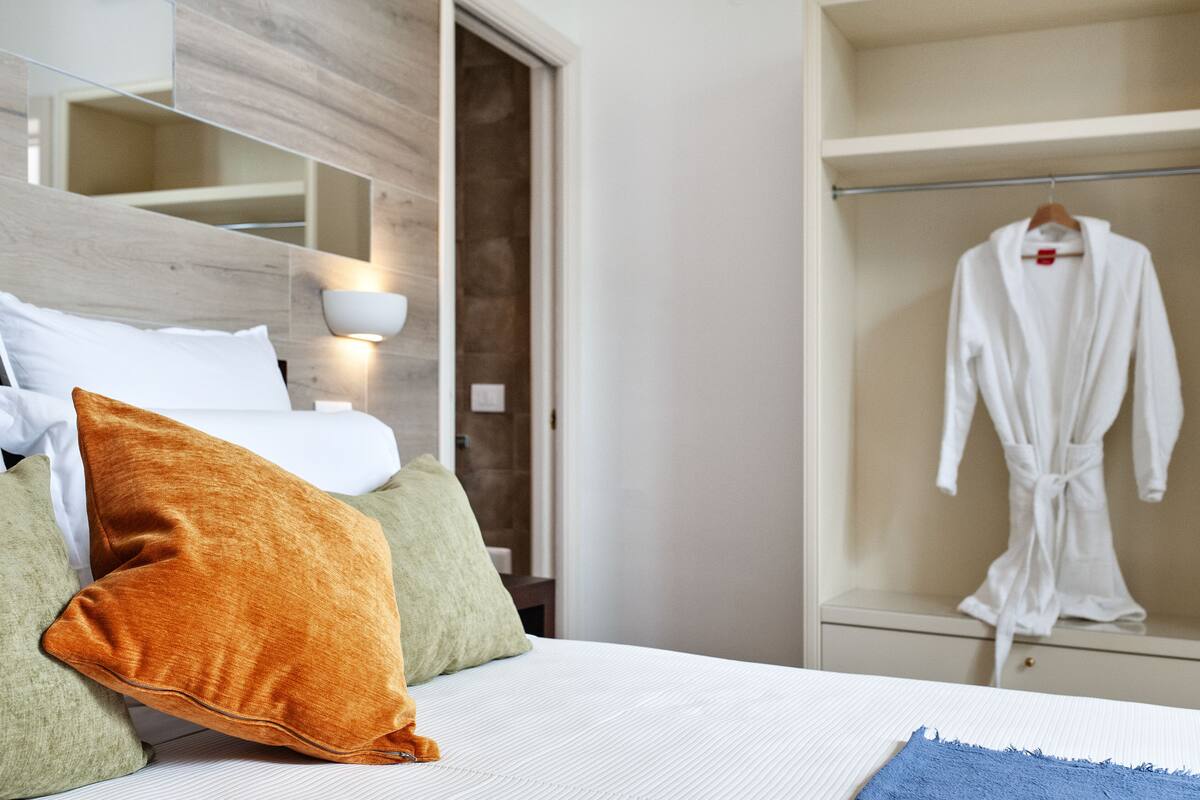 Ein Hotelzimmer mit Bett, Kissen, einem Bademantel, der an einem Kleiderständer hängt, und einem Nachttisch mit Lampe.
