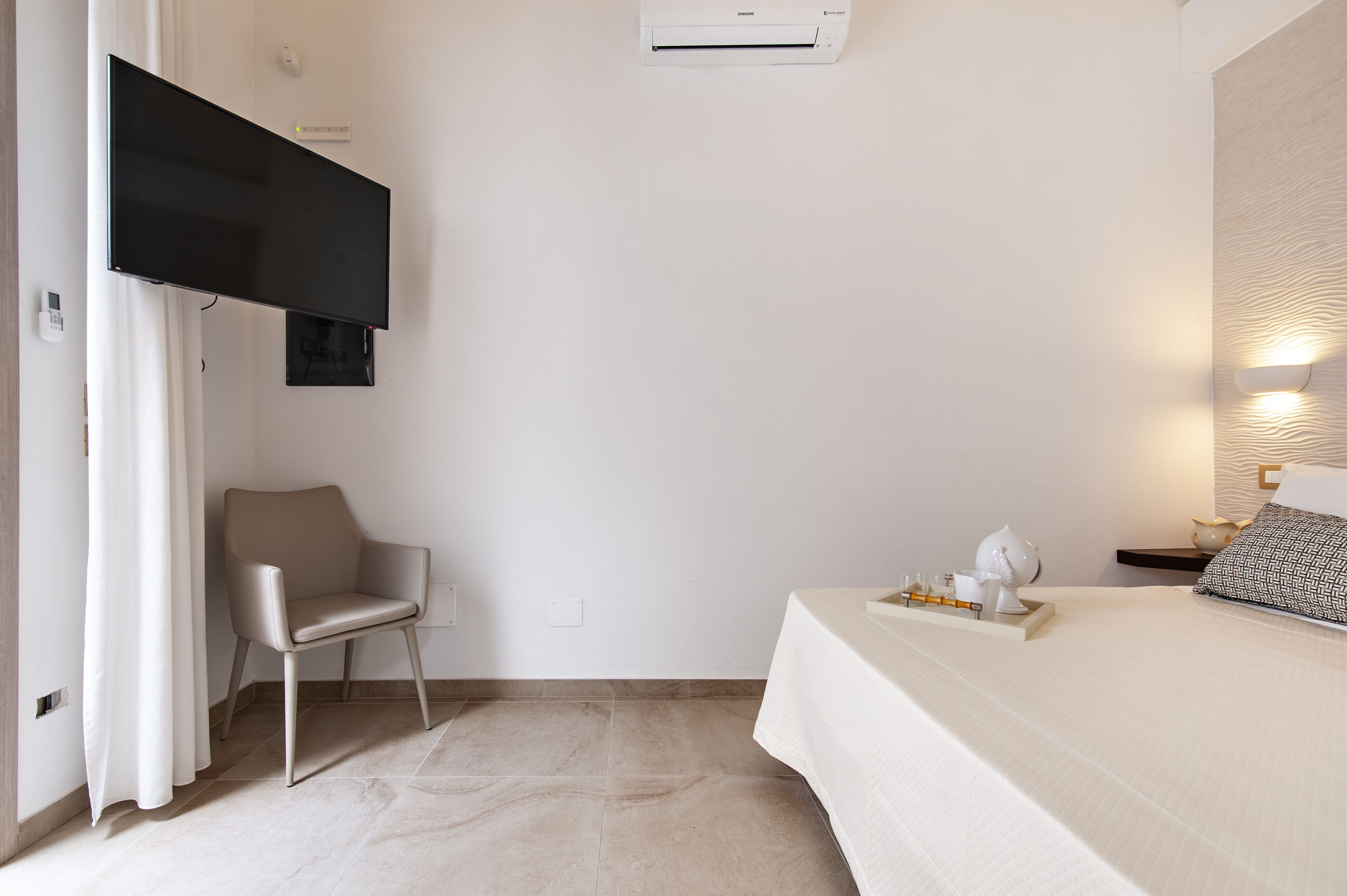 Une chambre d’hôtel moderne équipée d’un téléviseur à écran plat, d’un fauteuil, d’un lit avec un plateau contenant des articles de toilette et d’un climatiseur fixé au mur.