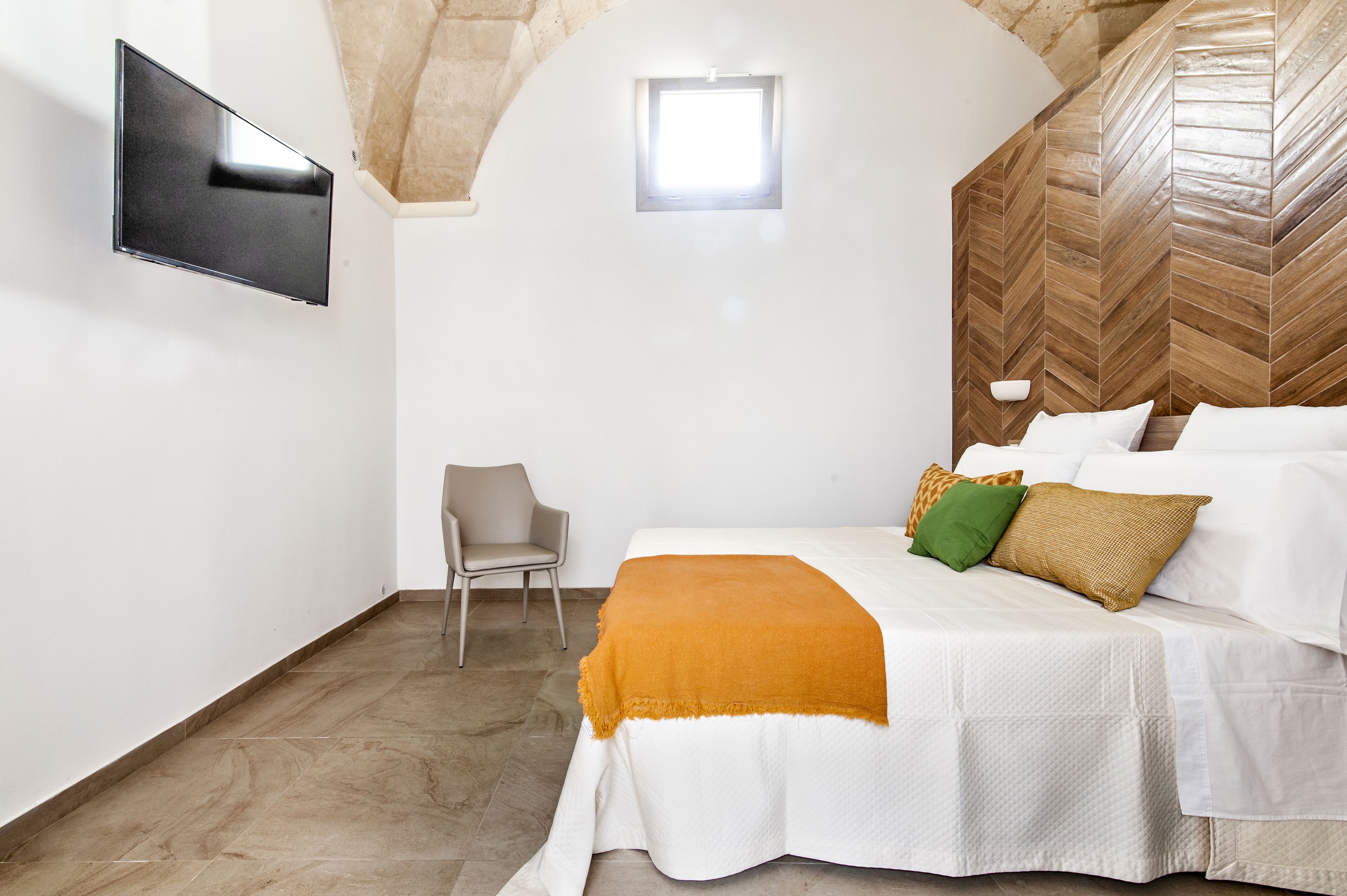 A bedroom with a bed, a chair, a TV mounted on the wall, and a window.
