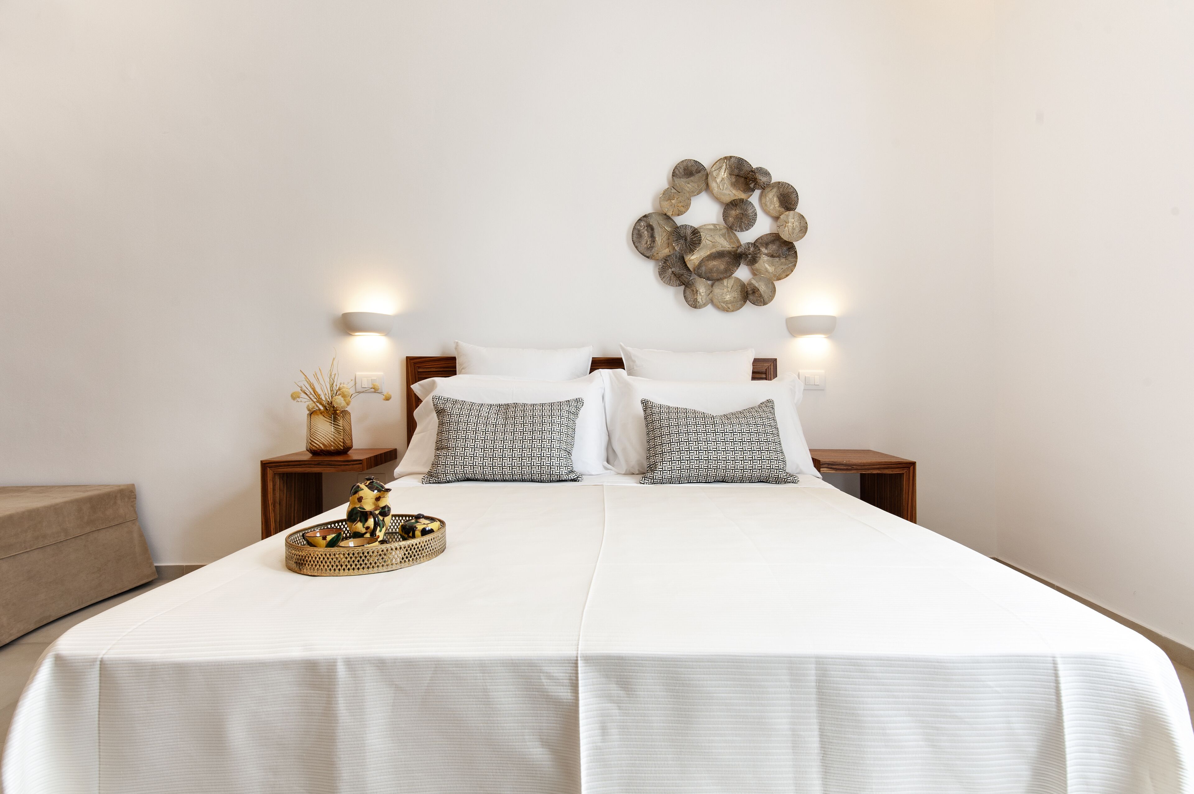 A neatly made bed with white bedding and patterned pillows, a decorative wall piece, and two bedside tables with lamps.