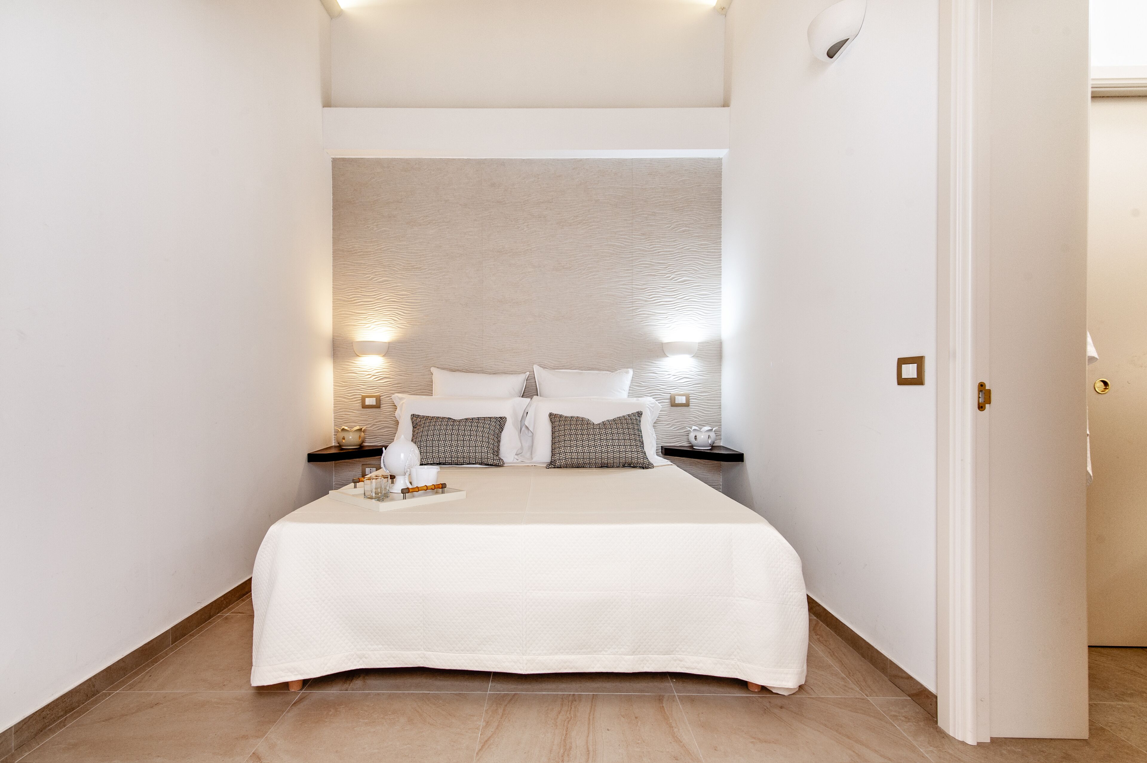 Un lit bien fait, recouvert d’une couvre-lit blanche et agrémenté d’oreillers, deux tables de chevet avec des lampes, et un mur à motifs derrière le lit.