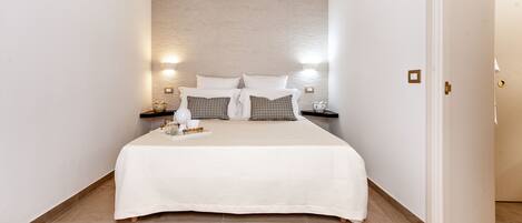 Un lit bien fait, recouvert d’une couvre-lit blanche et agrémenté d’oreillers, deux tables de chevet avec des lampes, et un mur à motifs derrière le lit.
