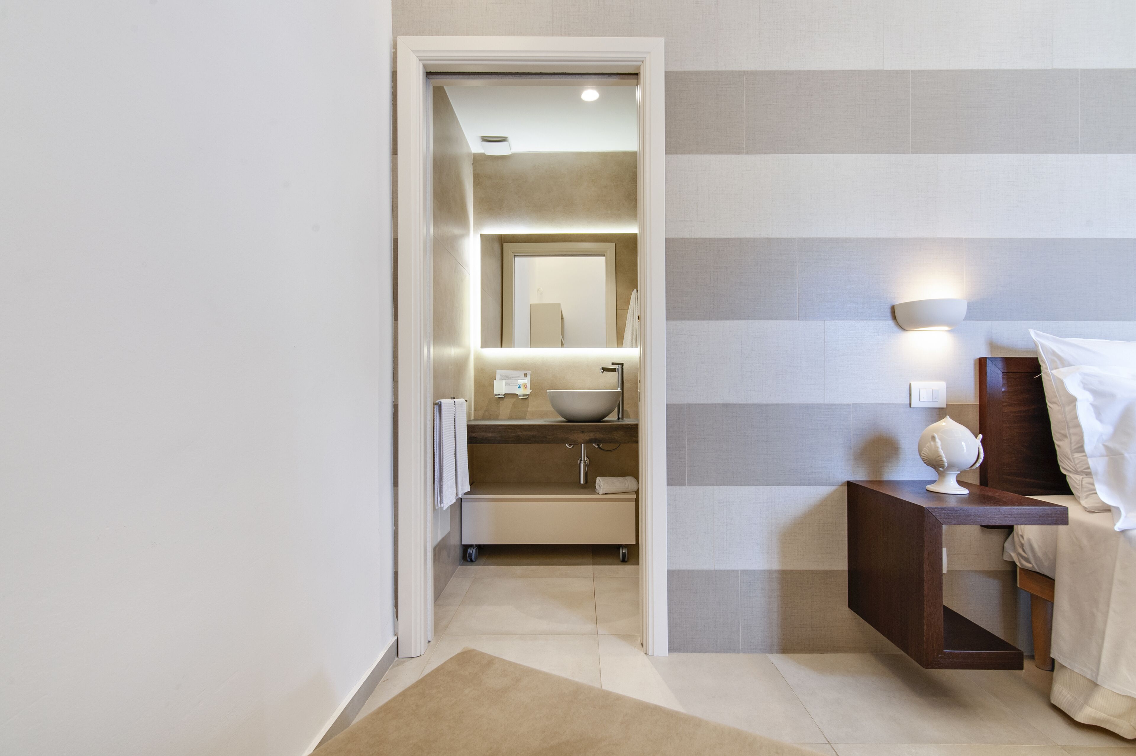 A modern bathroom with a large mirror, a bowl sink, and a bedside table with a lamp.