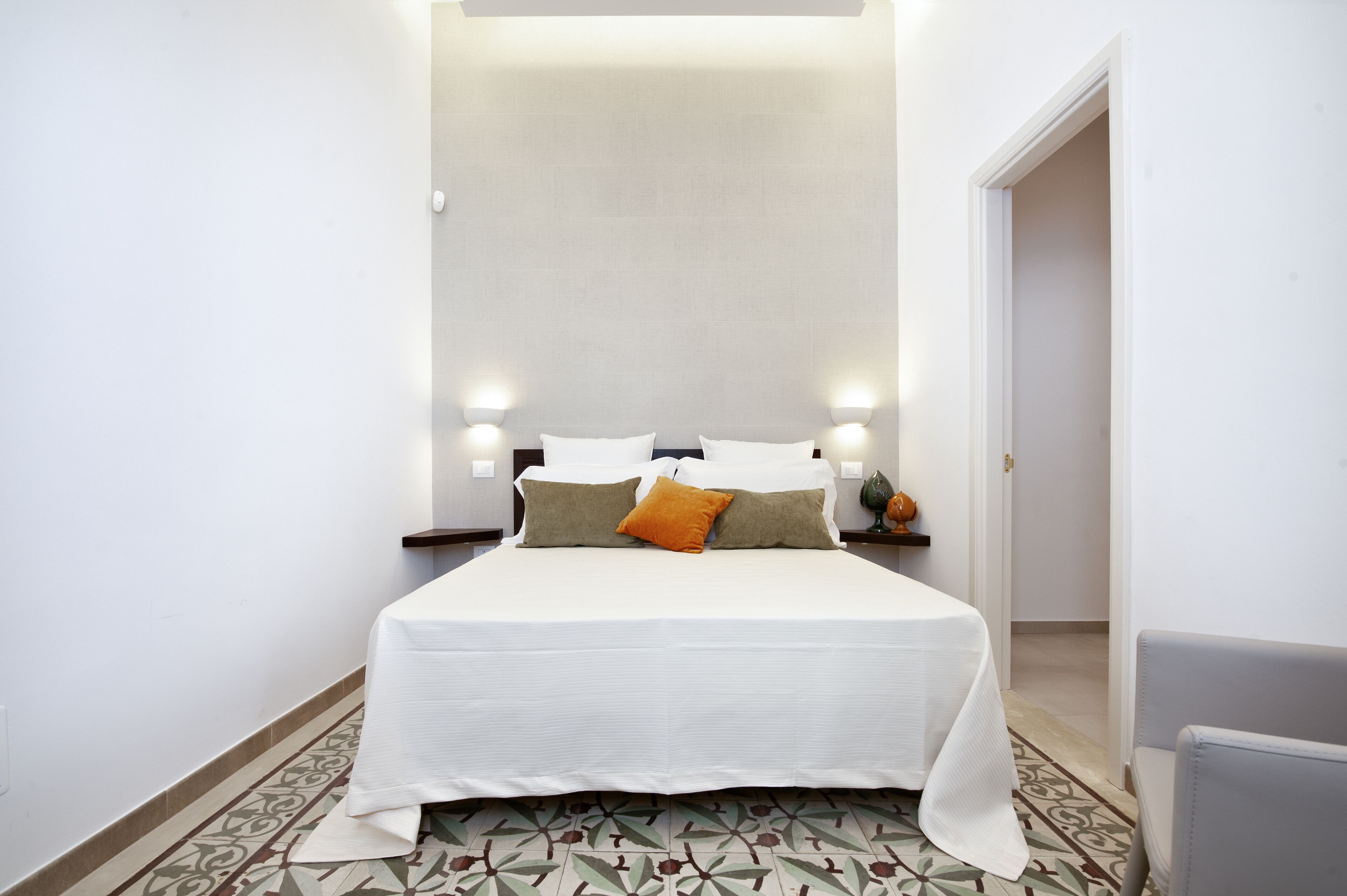 A neatly made bed with white bedding and two pillows, a patterned floor, and a small nightstand with a lamp.
