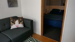 A room with a grey sofa, a cat-themed pillow, a bed with a blue blanket, and a wooden floor. - T1 Parking Inclus, Hyper Centre, Mais Très Calme (Pau)