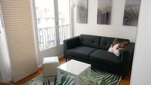 A modern living room with a dark grey sofa, a white coffee table, and a cat-themed pillow. There is a window with blinds, a white floor lamp, and two framed pictures on the wall. - T1 Parking Inclus, Hyper Centre, Mais Très Calme (Pau)