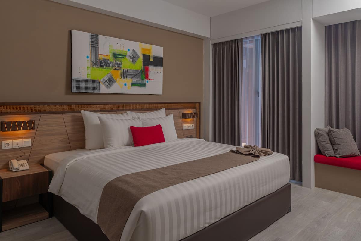 Ein modernes Hotelzimmer mit einem großen Bett, einem Nachttisch mit Telefon, einem Wandbild und Blick ins Freie durch ein Fenster mit Vorhängen.