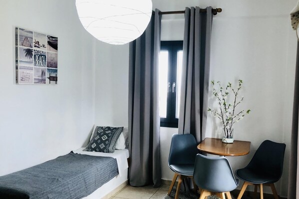 Quarto compacto com uma cama de solteiro, uma pequena mesa com duas cadeiras, uma janela com cortinas e um ponto de luz fixado na parede.