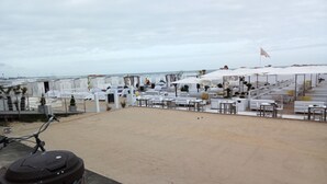 A beachside outdoor seating area with white tables and chairs, umbrellas, and a flag.