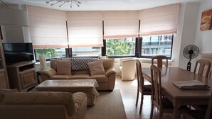 A modern living room with a sofa set, a TV, a dining table with chairs, and large windows with blinds.