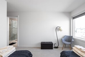 Une chambre moderne avec une douche en verre, un pouf noir, un fauteuil gris et une table de chevet avec des serviettes.