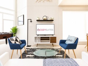 Uma sala de estar moderna com sofá, mesa de centro e uma televisão de tela plana exibindo o logotipo 'COZY SUITES'.