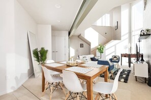 Un espace de repas moderne et bien éclairé, avec une table en bois et des chaises blanches, un escalier menant à un niveau supérieur et un coin salon confortable avec un canapé et des fauteuils.