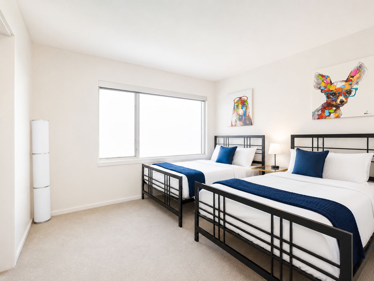 A hotel room with two beds, a large window, a trash can, and artwork on the wall.