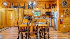 Interno di una baita in legno con un tavolo da pranzo centrale, sedie in legno, un lampadario e elettrodomestici da cucina.