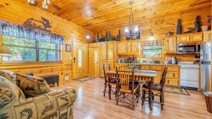 Un accogliente interno di una baita con pareti e pavimenti in legno, una zona pranzo con tavolo e sedie, una cucina con elettrodomestici moderni e un'ampia finestra vista alberi.