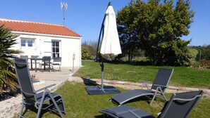 Une terrasse avec deux chaises longues et un parasol blanc, un bâtiment blanc avec un toit rouge, et une zone herbeuse avec des arbres.
