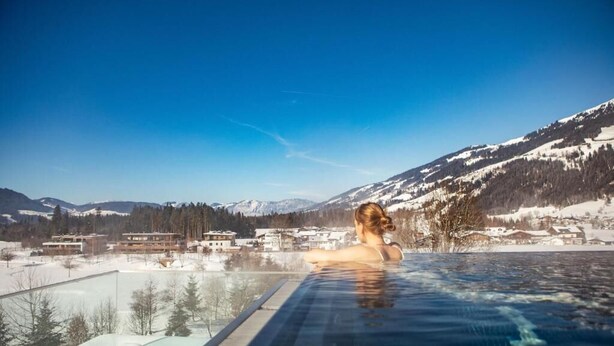 Une personne se trouve dans un bain à remous, d’où elle peut admirer un paysage montagneux enneigé, avec des bâtiments visibles au loin.
