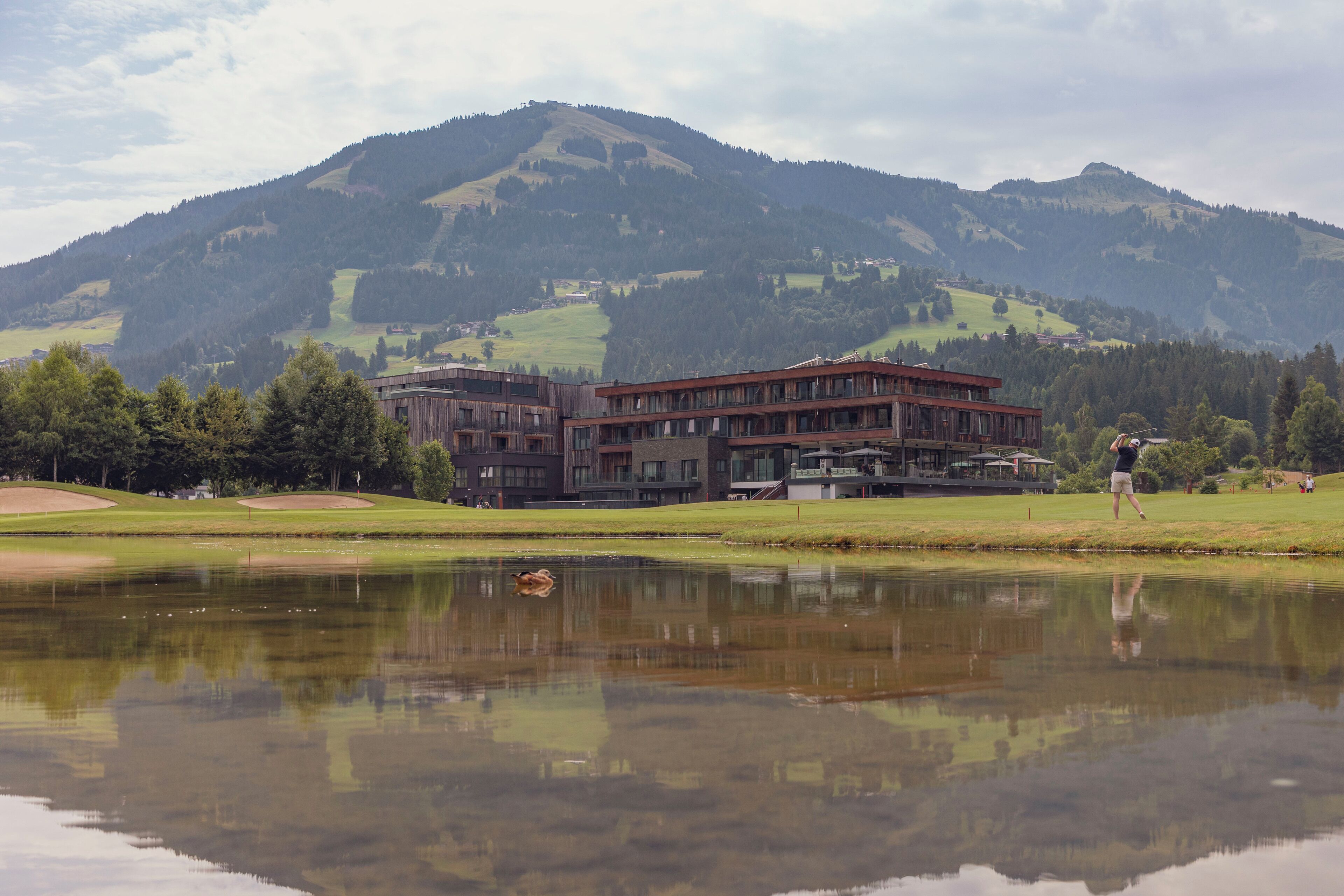 A hotel with a reflection in a pond, surrounded by a golf course and mountains.