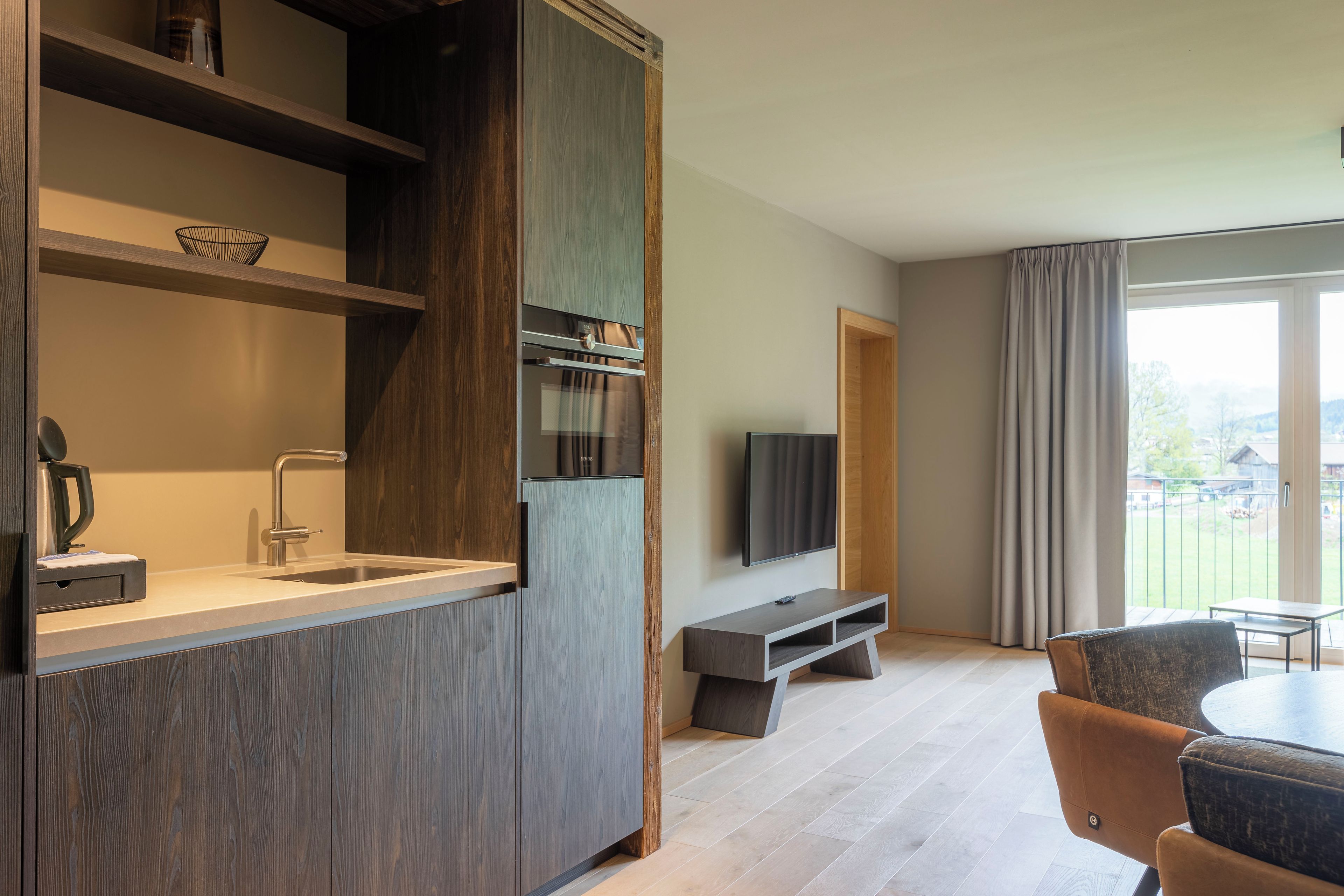 A modern kitchen with a built-in oven and sink, a flat-screen TV mounted on the wall, and a dining area with a table and chairs.