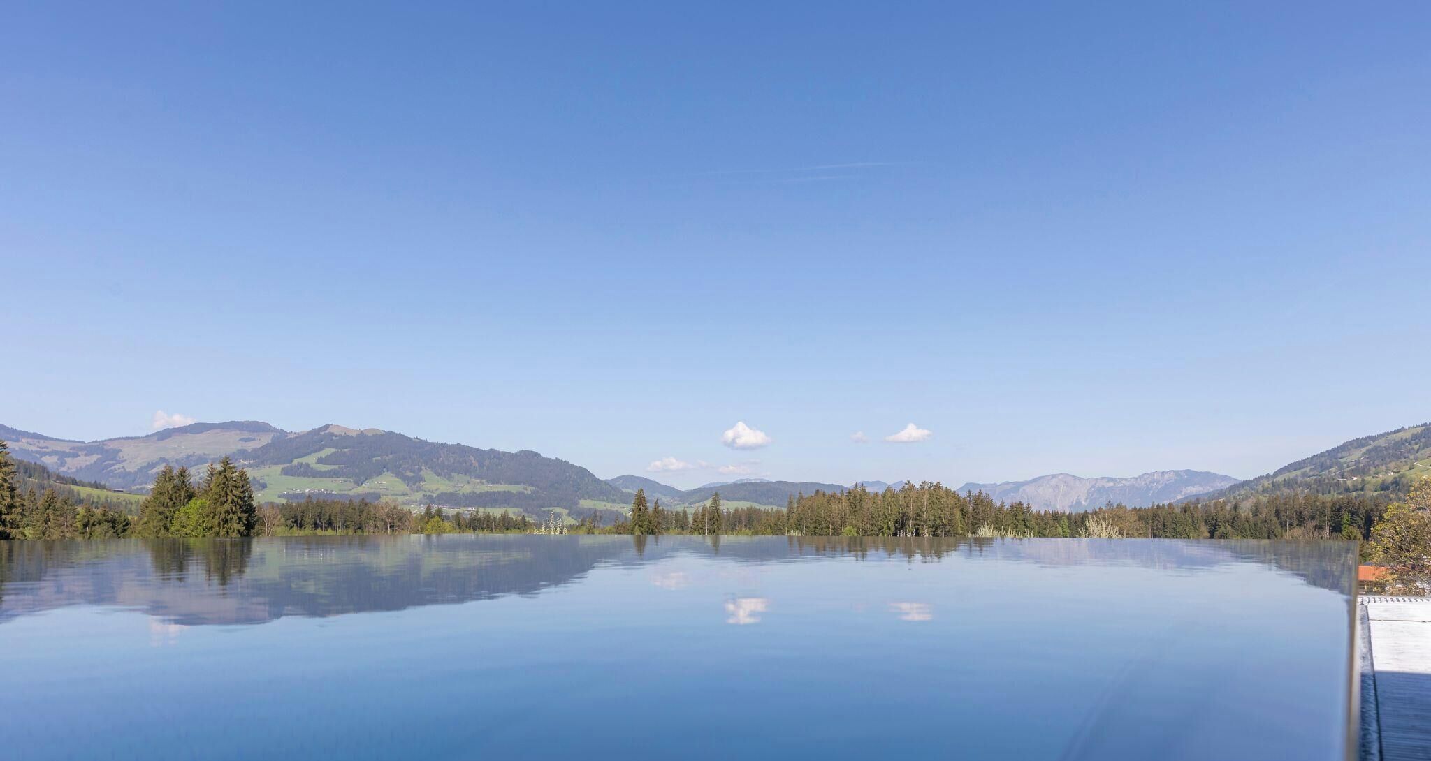 A calm lake with clear reflections, surrounded by forested hills and mountains under a clear sky.