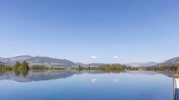 Ein ruhiger See mit klaren Spiegelungen, umgeben von bewaldeten Hügeln und Bergen unter blauem Himmel.