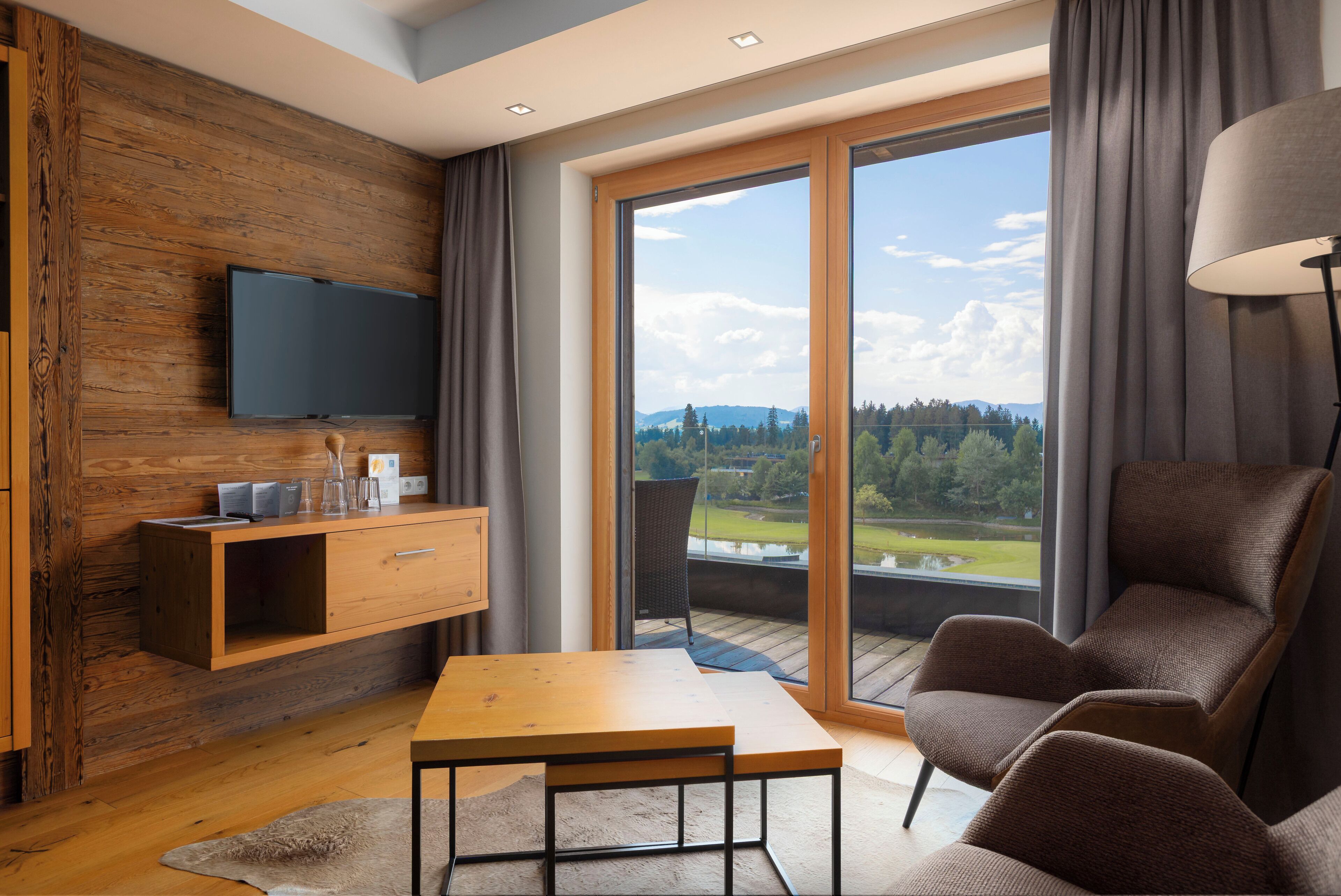 Una sala de estar moderna con una pared de madera, un televisor de pantalla plana, una mesa de madera y un amplio ventanal corredizo que da a un balcón con vistas a un lago y árboles.