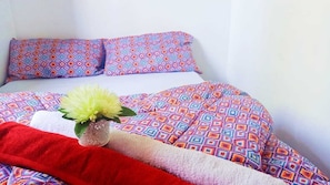 A bed with patterned bedding, a red blanket, and a decorative vase with flowers. - Cosmos Masawa (Homa Bay)
