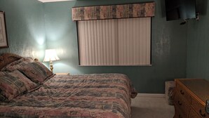 Un dormitorio con cama, cómoda, ventana con persianas, lámpara y televisión en la pared.