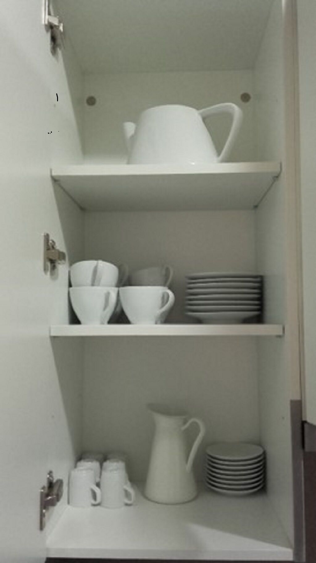 Un mueble blanco con tres estantes. El estante superior sostiene una jarra blanca y un tazón blanco. El estante central contiene tazas blancas y una pila de platos blancos. El estante inferior tiene una jarra blanca, una pila de platos blancos y una taza blanca.