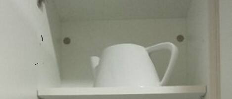 Un mueble blanco con tres estantes. El estante superior sostiene una jarra blanca y un tazón blanco. El estante central contiene tazas blancas y una pila de platos blancos. El estante inferior tiene una jarra blanca, una pila de platos blancos y una taza blanca.