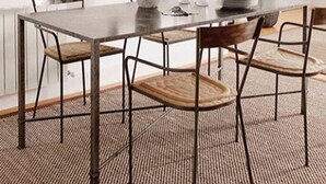 Uma sala de jantar com uma mesa retangular, quatro cadeiras, um vaso com flores e uma janela grande com moldura decorativa.