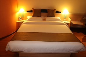 Una cama bien hecha con ropa de cama blanca y marrón, dos lámparas de noche y una mesita de noche con un teléfono y un libro.