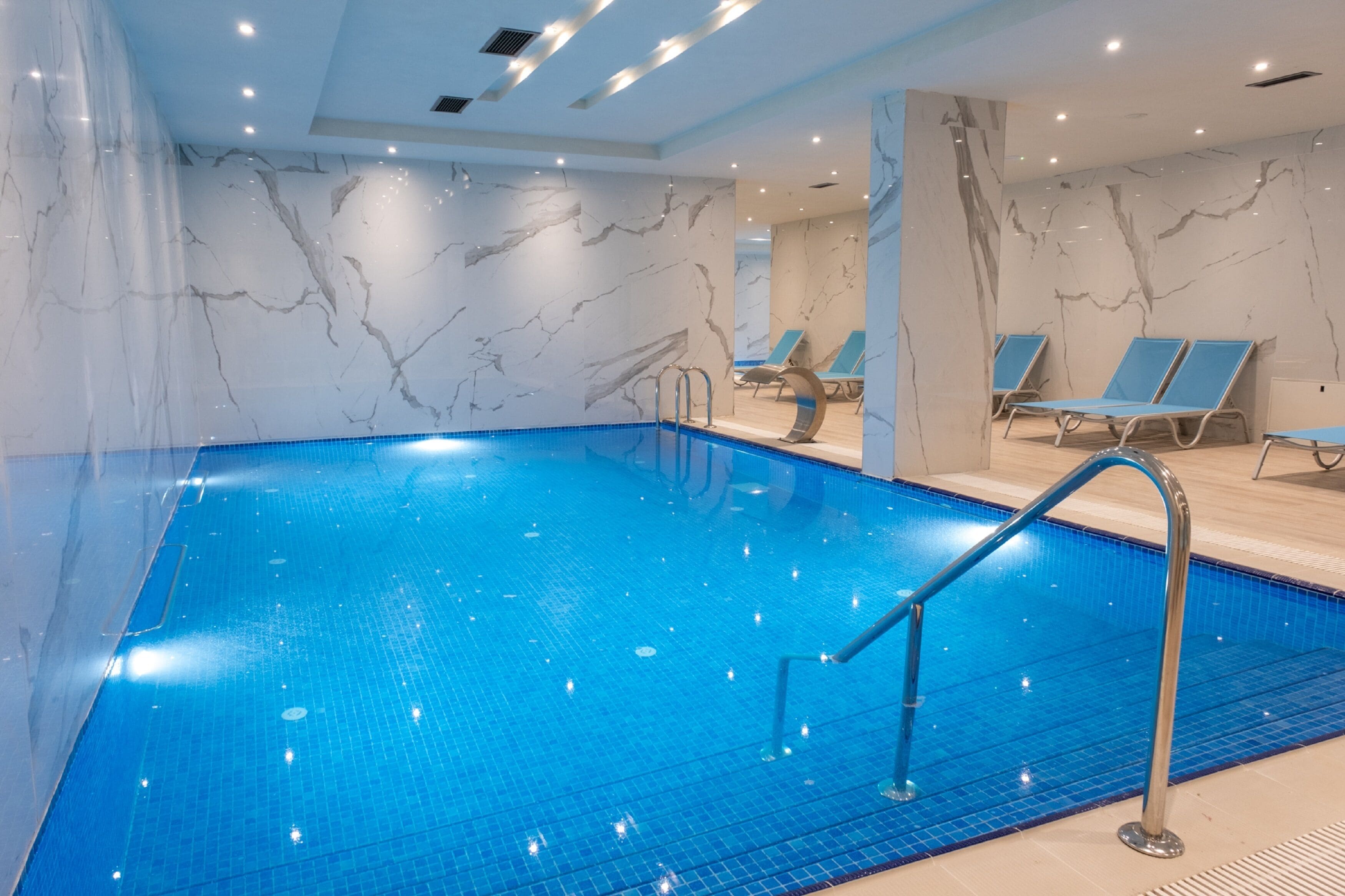 A swimming pool with blue tiles, a marble wall, and lounge chairs.
